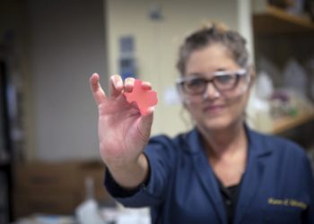 Woman In Lab Holding Small Texas Shaped Item In Hand Toward Camera