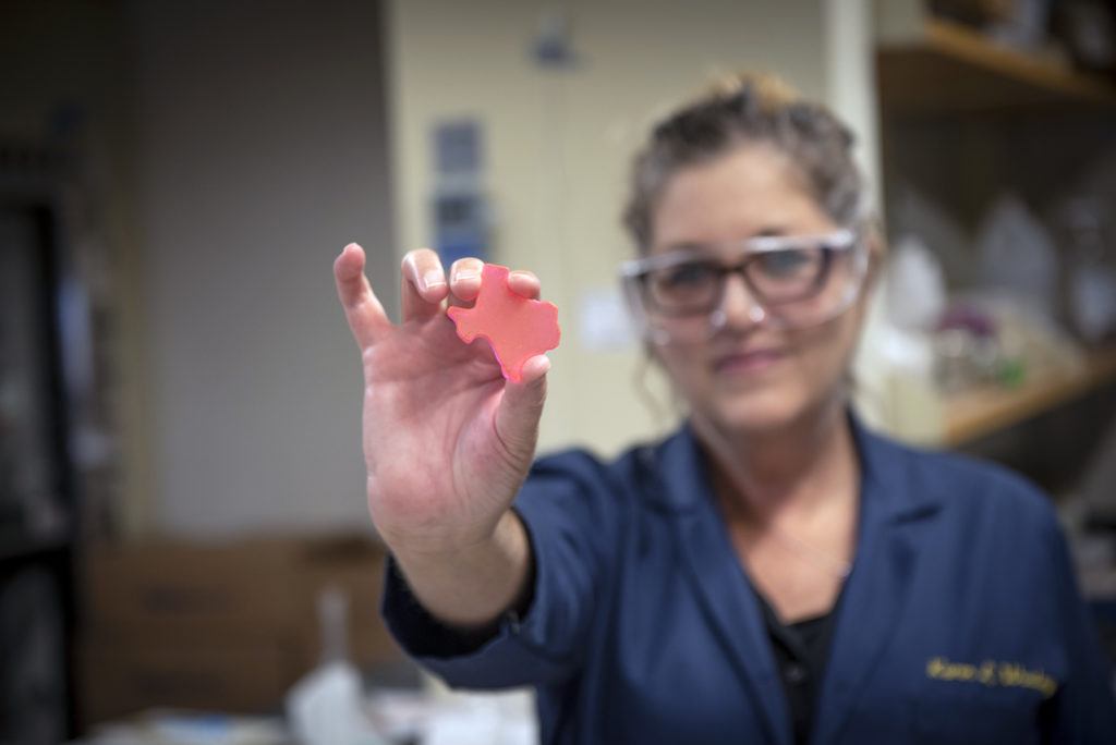 woman in lab holding small texas shaped item in hand toward camera