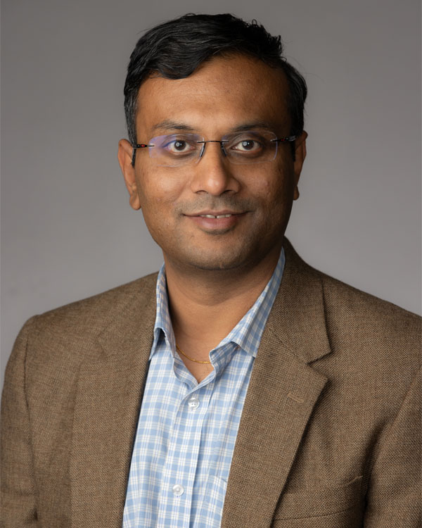 Man in a blue and white checkered shirt, brown blazer, and wearing glasses